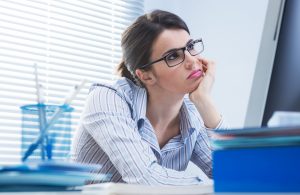 Bored office worker at desk staring at computer screen with hand on chin.