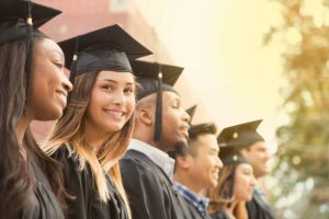 Latin descent girl waits in line during college graduation ceremony. She looks at camera with a big smile as she wears a black cap and gown.